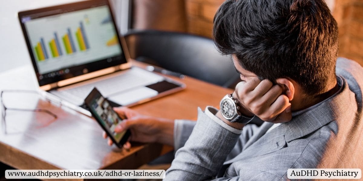 Man Scrolling on Phone in front of work computer, representing differences between ADHD paralysis vs procrastination 